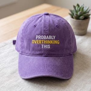 May include: A purple baseball cap featuring the embroidered text PROBABLY OVERTHINKING THIS in white and yellow lettering. The cap has a structured crown, a curved brim, and a button on top, with a classic six-panel design and stitched eyelets.