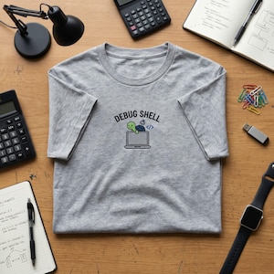 May include: A grey t-shirt with a crew neck. The shirt displays the text "DEBUG SHELL" above a graphic of a turtle on a laptop screen with a code symbol. The shirt is on a wooden surface with office supplies.