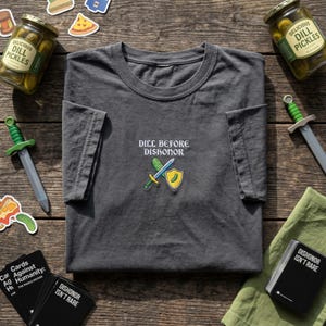 May include: Charcoal grey t-shirt featuring the embroidered text "DILL BEFORE DISHONOR" and a cartoon pickle sword and shield. The image also shows jars of pickles, toy swords, and a card game.