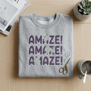 Peut inclure: Un sweat-shirt gris avec le mot "AMAZE!" en violet, répété trois fois. La lettre "Z" de chaque mot est ornée d'un motif de feuille verte. Une paire de ciseaux dorés et une tasse de café sont à côté du sweat-shirt.