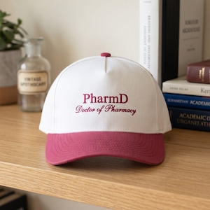 May include: A white baseball cap with a claret brim and button. The cap features the embroidered text "PharmD Doctor of Pharmacy" in claret. The hat is displayed on a wooden surface.