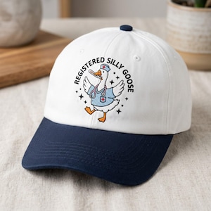 May include: A white and navy blue baseball cap with the text "REGISTERED SILLY GOOSE" and an embroidered cartoon goose wearing a nurse's uniform. The cap has a curved brim and a button on top.