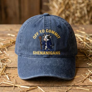 May include: A navy blue baseball cap with the embroidered phrase "OFF TO COMMIT SHENANIGANS." The cap features a cartoon cow character in a robe holding a staff. The hat is made of a soft, worn material.
