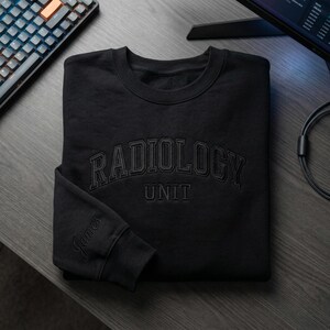 May include: Black crew neck sweatshirt featuring the embroidered text "RADIOLOGY UNIT". The name "James" is embroidered on the left sleeve. The sweatshirt is made of a soft material.