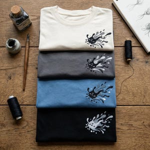 May include: Four folded t-shirts in cream, gray, blue, and black, each with an embroidered abstract design in black, gray, and white. The shirts are stacked on a wooden surface, with ink and art supplies nearby.