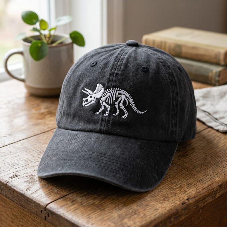 May include: A black baseball cap with a curved brim. The cap features a white embroidered design of a triceratops skeleton on the front. The hat is made of a soft, worn material and has a top button.