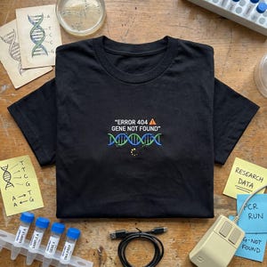 May include: A black short-sleeved t-shirt featuring an embroidered graphic of a DNA double helix in blue and green, with the text "ERROR 404 GENE NOT FOUND" in white, an orange warning triangle icon, and a yellow circular loading symbol.