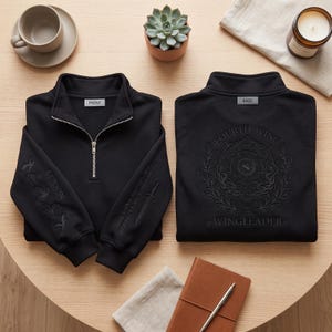 May include: Two black quarter-zip sweatshirts. The left one shows the front with a silver zipper, "RIORSON" text, and a sword graphic on the sleeves. The right one shows the back with an embossed "FOURTH WING WINGLEADER" circular design.