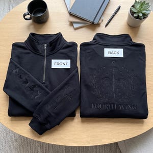 May include: Two black quarter-zip sweatshirts. The left one features a sword and dragon embroidery on the sleeve with the text 'Violent Little Thing'. The right one displays a large dragon, laurel, and 'FOURTH WING' text on the back.