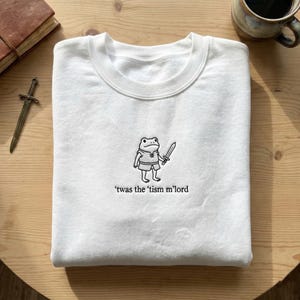 May include: White crewneck sweatshirt featuring a black and white embroidered frog holding a sword. The phrase "'twas the 'tism m'lord" is embroidered below the frog. The sweatshirt is on a wooden surface.