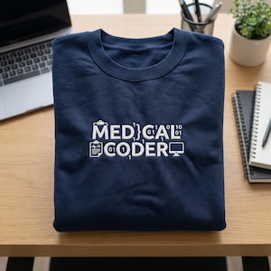 May include: A navy blue sweatshirt featuring the white text "MEDICAL CODER" with binary code elements and medical-themed icons. The garment is folded on a wooden surface, alongside a laptop and stationery.