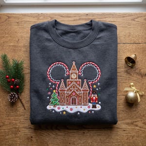 Peut inclure: Un sweat-shirt gris foncé à col rond orné d'une broderie représentant un château en pain d'épices au centre d'un contour en sucre d'orge. Le motif inclut un petit sapin de Noël, un casse-noisette et des détails de glaçage blanc.