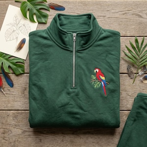 Peut inclure: Un sweat-shirt vert foncé à fermeture éclair quart de tour, orné d'une broderie représentant un perroquet rouge, jaune et bleu perché sur une branche avec des feuilles vertes. Le vêtement possède une fermeture éclair métallique argentée.