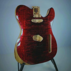 May include: An electric guitar body with a deep red, high-gloss finish over a visible wood grain pattern. The sides and neck pocket feature natural light-coloured wood. It includes routed cavities for pickups and bridge, and a logo stamp.