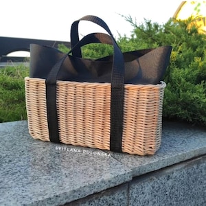 May include: A rectangular woven basket with black fabric lining and black handles. The basket is made of light brown wicker and has a sturdy construction. The text "SVITLANA POLONSKA" is printed on the front of the basket.