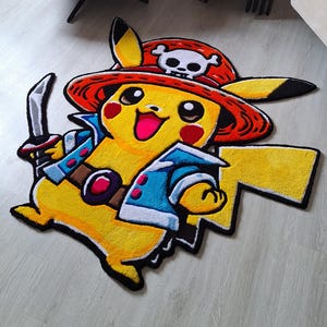 K&ouml;nnte beinhalten: Ein getufteter Teppich in Form der Figur Pikachu, die einen roten Piratenhut mit einem wei&szlig;en Totenkopf-Emblem, eine blaue Jacke tr&auml;gt und ein silbernes Schwert h&auml;lt. Die Figur ist gelb mit schwarzen Ohrspitzen, roten Wangen und einem Blitzschwanz.