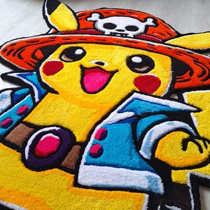 K&ouml;nnte beinhalten: Ein getufteter Teppich in Form der Figur Pikachu, die einen roten Strohhut mit einem wei&szlig;en Totenkopf-Emblem tr&auml;gt. Die Figur hat gelbes Fell, rote Wangen, eine blaue Jacke mit wei&szlig;em Saum und einen braunen G&uuml;rtel mit violetter Schnalle.