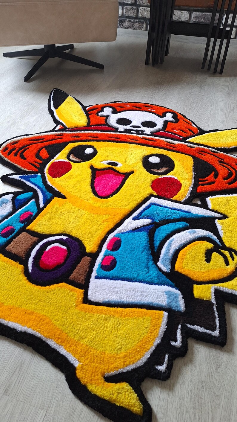 K&ouml;nnte beinhalten: Ein getufteter Teppich in Form der Figur Pikachu, die einen roten Strohhut mit einem wei&szlig;en Totenkopf-Emblem tr&auml;gt. Die Figur hat gelbes Fell, rote Wangen, eine blaue Jacke mit wei&szlig;em Saum und einen braunen G&uuml;rtel mit violetter Schnalle.