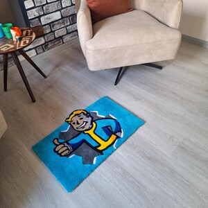 May include: A blue rug with a cartoon character from the Fallout video game series. The character is wearing a blue jumpsuit and a yellow utility belt. The character is giving a thumbs up. The rug is on a light wood floor.