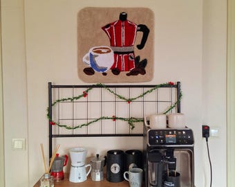 May include: A black coffee maker with a silver drip tray and a red and black coffee pot rug hanging on the wall. There are several coffee pots and mugs on a wooden shelf below the coffee maker.