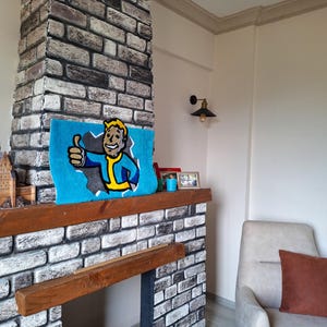 May include: A brick fireplace with a wooden mantel and a blue rug with a cartoon character on it. The rug has the text "Vault-Tec" on it. There is a small armchair with a brown pillow in the corner of the room.