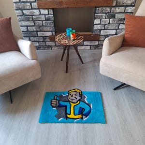 May include: A blue rug with a cartoon character from the video game Fallout. The character is wearing a blue jumpsuit and has a yellow collar. The character is giving a thumbs up. The rug is on a light brown floor in front of a white couch and a wooden coffee table.