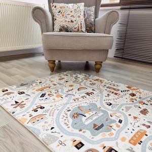 Puede incluir: Una alfombra de juego para ni&ntilde;os con un dise&ntilde;o de mapa de carreteras que presenta coches, trenes y barcos en tonos azules, naranjas y marrones. Dos cojines a juego descansan sobre un sill&oacute;n beige. La alfombra es rectangular.