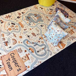Puede incluir: Una alfombra de juego con un dise&ntilde;o de mapa de carreteras, con carreteras, edificios y veh&iacute;culos en tonos beige, marr&oacute;n y azul. La alfombra tiene un letrero de madera que dice "Cooper's Town". Varias almohadas estampadas est&aacute;n apiladas sobre la alfombra.