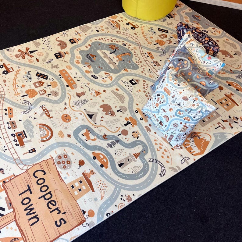 Puede incluir: Una alfombra de juego con un dise&ntilde;o de mapa de carreteras, con carreteras, edificios y veh&iacute;culos en tonos beige, marr&oacute;n y azul. La alfombra tiene un letrero de madera que dice "Cooper's Town". Varias almohadas estampadas est&aacute;n apiladas sobre la alfombra.
