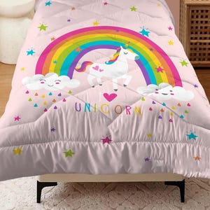 Pode incluir: Uma colcha rosa com um arco-íris, um unicórnio branco e o texto "I Unicorn". A colcha tem um padrão acolchoado e é decorada com estrelas e gotas de chuva coloridas. O design é caprichoso e divertido.