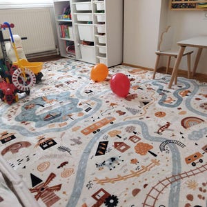Puede incluir: Una alfombra de juego para ni&ntilde;os con un dise&ntilde;o de carretera y ciudad en colores neutros. La alfombra presenta carreteras, edificios, trenes y otras ilustraciones. Dos globos, uno naranja y otro rojo, est&aacute;n sobre la alfombra. Varios veh&iacute;culos de juguete est&aacute;n cerca.