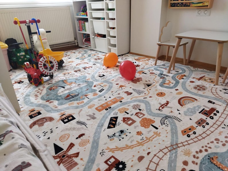 Puede incluir: Una alfombra de juego para ni&ntilde;os con un dise&ntilde;o de carretera y ciudad en colores neutros. La alfombra presenta carreteras, edificios, trenes y otras ilustraciones. Dos globos, uno naranja y otro rojo, est&aacute;n sobre la alfombra. Varios veh&iacute;culos de juguete est&aacute;n cerca.