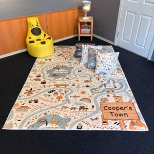 Puede incluir: Una alfombra de juego beige con un dise&ntilde;o de mapa de carreteras, con carreteras, r&iacute;os e ilustraciones de animales, veh&iacute;culos y edificios. La alfombra tiene un letrero que dice "Cooper's Town". Tambi&eacute;n se ven varias almohadas decorativas y un puf amarillo.