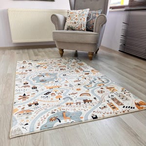 Puede incluir: Una alfombra de juego rectangular con un dise&ntilde;o de mapa de carreteras. La alfombra presenta carreteras, edificios, veh&iacute;culos y animales en tonos de azul, naranja y marr&oacute;n sobre un fondo blanco. Un sill&oacute;n gris y cojines decorativos est&aacute;n en el fondo.