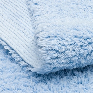 May include: A close-up of a light blue bath mat with a fluffy texture. The mat is folded in half, showing the different textures of the fabric.