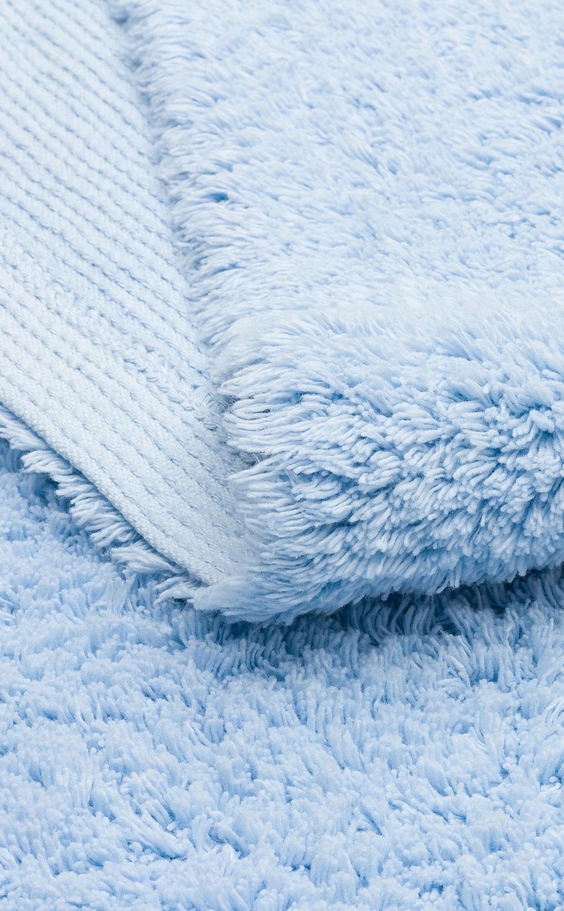 May include: A close-up of a light blue bath mat with a fluffy texture. The mat is folded in half, showing the different textures of the fabric.