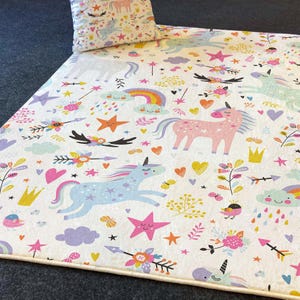 Puede incluir: Una alfombra de juego blanca con un patrón colorido de unicornio, arcoíris, estrellas y corazones. La alfombra de juego tiene un borde marrón claro.