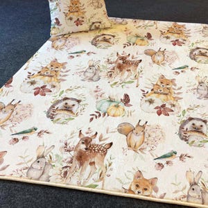 Puede incluir: Una alfombra de color crema con estampado de animales del bosque. La alfombra presenta una variedad de animales, incluyendo ciervos, zorros, ardillas, erizos y conejos. Los animales están representados en un estilo acuarela y están rodeados de hojas y flores.