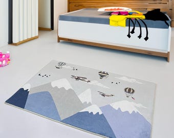Puede incluir: Una alfombra azul y gris con un diseño de montaña y avión. La alfombra presenta globos aerostáticos y aviones volando sobre las montañas.