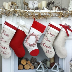 Op de afbeelding: Vier kerstsokken hangen aan een witte schouw. De sokken zijn gemaakt van witte stof met rode bies en zijn geborduurd met de woorden "Cats Love Santa Too!" en "Dogs Love Santa Too!" Eén sok heeft een rode strik en de tekst "Brandilee".