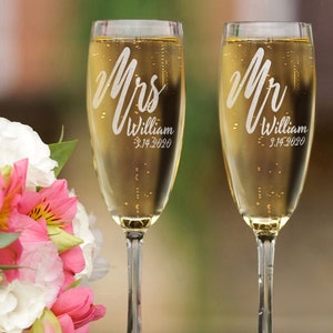 Pode incluir: Duas ta&ccedil;as de champanhe cheias de uma bebida borbulhante. As ta&ccedil;as s&atilde;o gravadas com "Mrs" e "Mr", um nome e uma data. Um buqu&ecirc; de flores brancas e rosas est&aacute; ao fundo.