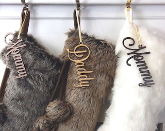 Op de afbeelding: Drie kerstsokken van nep bont met gepersonaliseerde houten naamschildjes. De sokken hangen aan haken boven een witte schouw. De sokken zijn bruin, bruin-wit en wit.