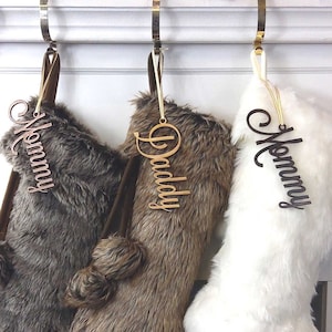 May include: Three faux fur Christmas stockings with personalized wooden name tags. The stockings are hung on hooks above a white fireplace mantel. The stockings are brown, brown and white, and white.