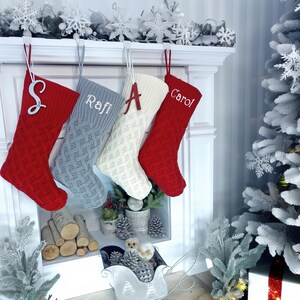 May include: Four cable-knit Christmas stockings in red, gray, and cream hang from a white mantel. Each stocking is personalized with a letter or name: "S", "Rafi", "A", and "Carol". The stockings are decorated with a snowflake garland.