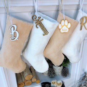 May include: Four plush Christmas stockings in cream and tan hues hang from a white mantel. Each stocking has a wooden tag with a name or word: "Bianca", "Mommy", "Kiwi", and "Richard". The "Kiwi" tag is a paw print.