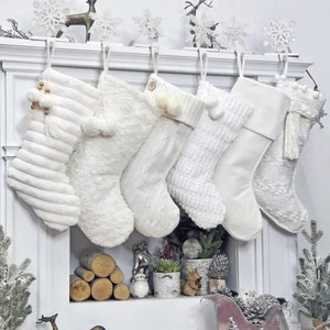 May include: A collection of six white Christmas stockings hanging from a white mantel. The stockings vary in texture, including ribbed, faux fur, and cable knit designs. Decorative elements include tassels, pom-poms, and wooden beads. The mantel is decorated with snowflakes and reindeer.