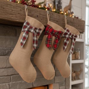 Peut inclure: Trois bas de Noël en toile de jute beige avec des nœuds écossais rouges et blancs suspendus à un manteau de cheminée en bois. Les bas sont suspendus par des crochets métalliques. La cheminée est en pierre grise.