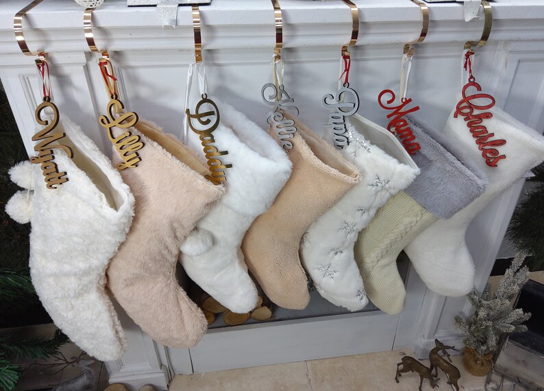 May include: Eight white, cream, and gray Christmas stockings with personalized wooden and acrylic name tags hanging on a white fireplace mantel. The names on the tags are Wyatt, Lilly, Daddy, Kellie, Prima, Mama, and Charles.