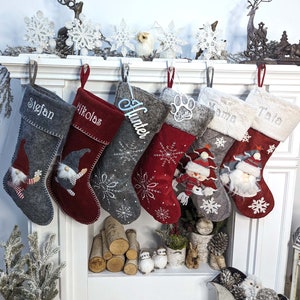 Puede incluir: Una colecci&oacute;n de calcetines navide&ntilde;os en tonos grises y rojos, cada uno personalizado con nombres como "Stefan" y "Nikolas". Algunos calcetines presentan dise&ntilde;os de gnomos y copos de nieve, mientras que otros tienen detalles de felpa y los nombres "Mama" y "Tata".