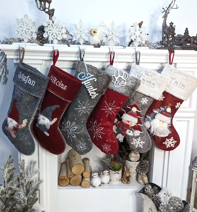 Puede incluir: Una colecci&oacute;n de calcetines navide&ntilde;os en tonos grises y rojos, cada uno personalizado con nombres como "Stefan" y "Nikolas". Algunos calcetines presentan dise&ntilde;os de gnomos y copos de nieve, mientras que otros tienen detalles de felpa y los nombres "Mama" y "Tata".
